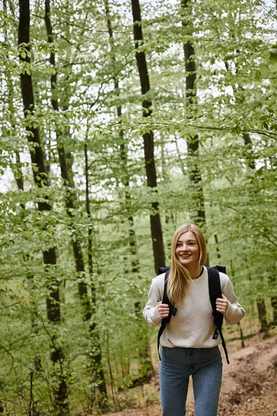 Sonriente senderismo mujer rubia con suéter y mochila caminando en el paisaje forestal en los bosques - foto de stock