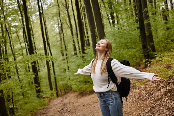 Smiling relaxed blonde woman hiker wearing sweater and backpack with arms  open in forest scenery — Stock Photo