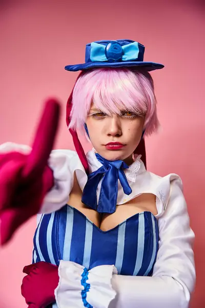 Attrayant cosplayer féminin en robe vibrante avec chapeau bleu pointant vers la caméra sur fond rose — Photo de stock