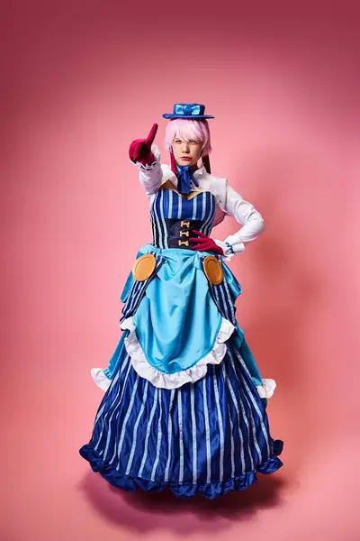 Alluring female cosplayer in vibrant dress with blue hat pointing at camera on pink backdrop — Stock Photo