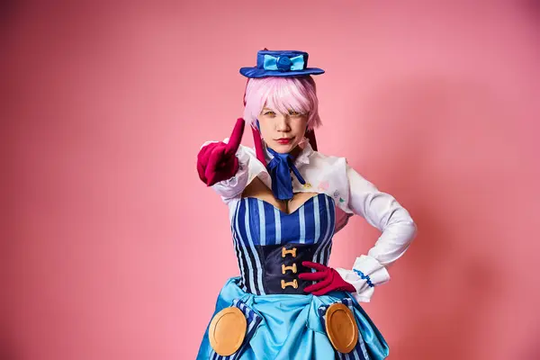 Alluring female cosplayer in vibrant dress with blue hat pointing at camera on pink backdrop — Stock Photo
