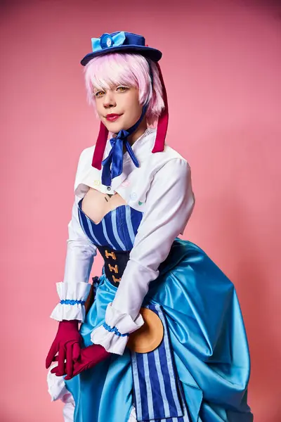 Cosplayer femenino sexy glamoroso en sombrero azul y traje vibrante mirando a la cámara en el telón de fondo rosa - foto de stock