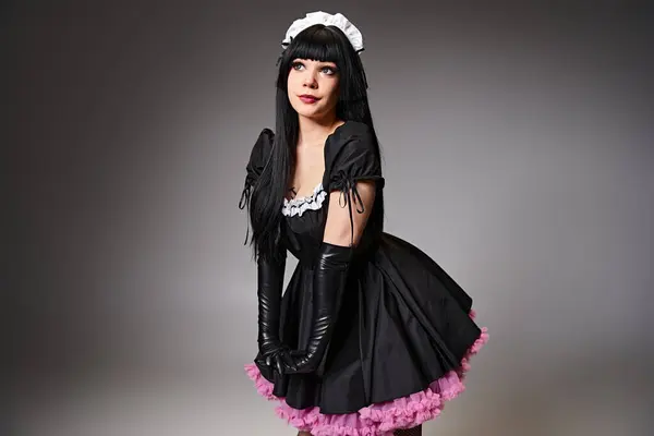 Attraente donna in costume da cameriera nera cosplaying personaggio anime e guardando lontano su sfondo grigio — Foto stock