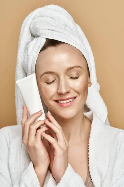 Una donna attraente con un asciugamano avvolto intorno alla testa tiene un tubo di crema, in mostra la sua naturale routine di bellezza. — Foto stock
