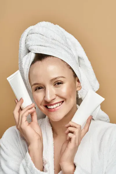 Eine Frau mit einem um den Kopf gewickelten Handtuch hält eine Tube Creme in der Hand und zeigt ihre Schönheitsroutine. — Stockfoto