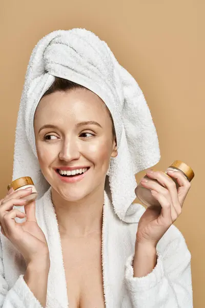 Una donna di bellezza naturale con un asciugamano in testa che tiene due barattoli di crema, indulgendo in un rituale di cura della pelle. — Foto stock