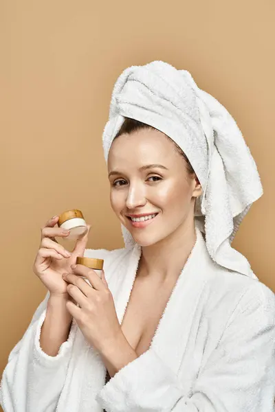 A stunning woman in a white robe holds cream with a look of contemplation and indulgence. — Stock Photo
