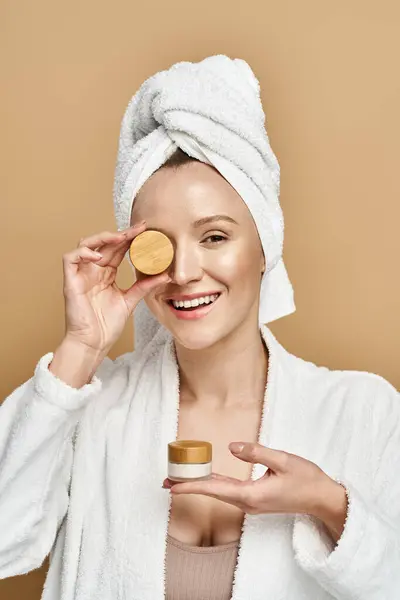 An attractive woman with a towel on her head holds a jar of cream, showcasing her natural beauty routine. — Stock Photo