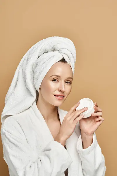 Una mujer serena con una toalla en la cabeza sosteniendo una crema en sus manos. - foto de stock