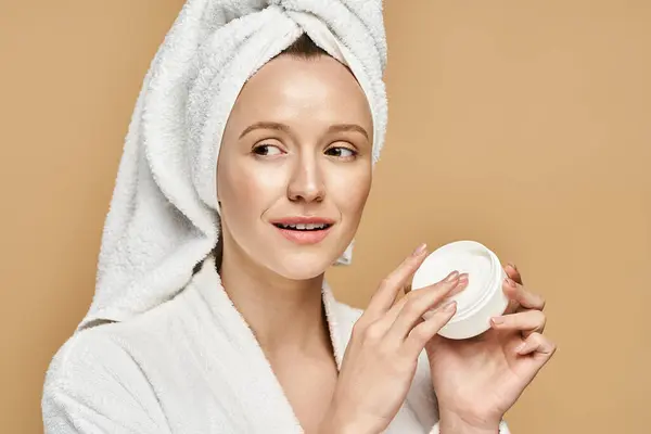 Una mujer de belleza natural sostiene un frasco de crema con una toalla en la cabeza. - foto de stock