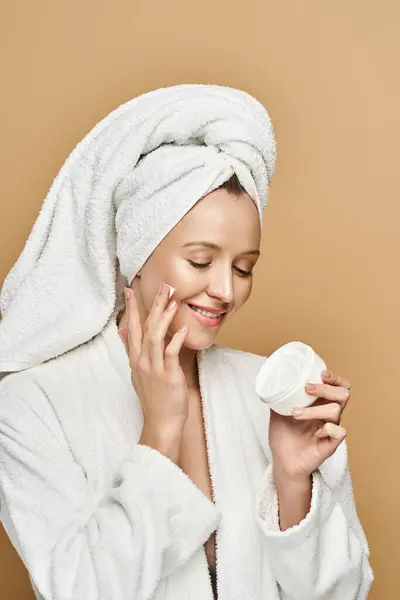 Uma mulher bonita com uma toalha enrolada em torno de sua cabeça segura um frasco de creme, exalando beleza natural e elegância. — Fotografia de Stock
