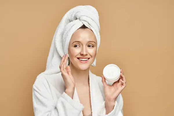 Une femme avec une serviette sur la tête tient un pot de crème, mettant en valeur sa routine beauté dans un cadre serein. — Photo de stock