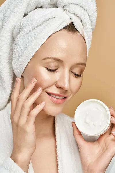 Une femme magnifique avec une serviette enroulée autour de sa tête est vue tenant un pot de crème, prêt à l'appliquer sur sa peau. — Photo de stock