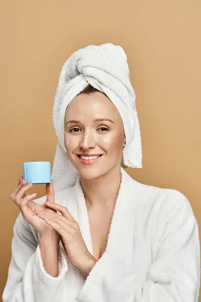 Una donna attraente con un asciugamano in testa regge con grazia vaso di crema, trasudando bellezza naturale e tranquillità.. — Foto stock
