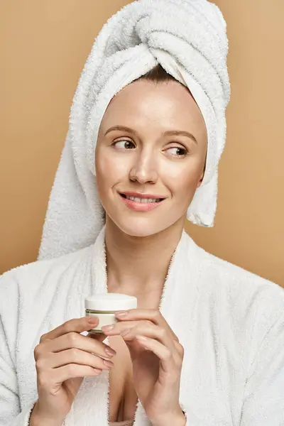 A natural beauty smirking glamorously, with a towel wrapped around her head, holding jar of cream close to her heart. — Stock Photo