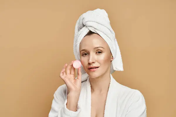 Una mujer con una toalla en la cabeza sostiene juguetonamente la crema, exudando belleza natural y gracia. - foto de stock