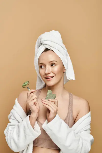 Женщина с полотенцем, обернутым вокруг головы, демонстрирующая естественную красоту, нежно держит в руке ролик для лица. — стоковое фото