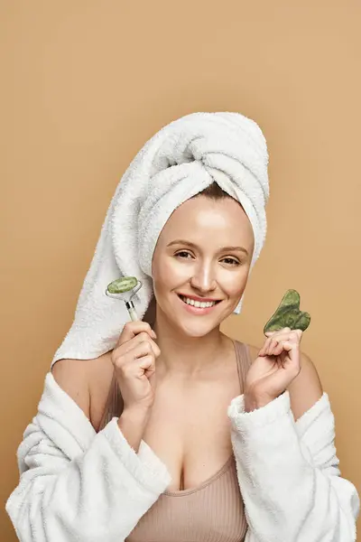 Une femme exsudant la beauté naturelle porte un turban de serviette tout en tenant délicatement un rouleau de visage dans une pose sereine. — Photo de stock