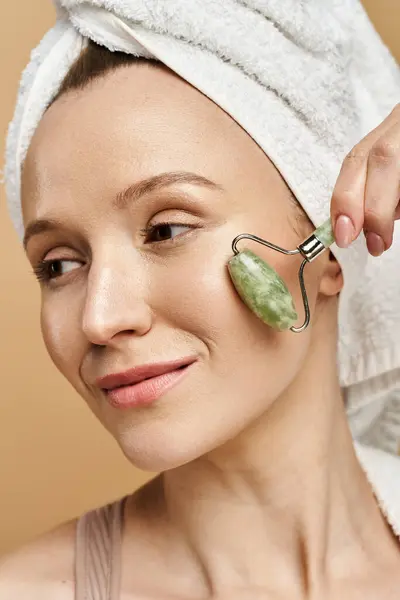 A woman with a towel on her head indulges in self-care with a face roller, radiating tranquility and natural beauty. — Stock Photo