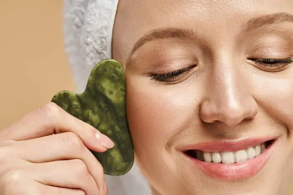 A serene and attractive woman with a towel wrapped around her head holds a vibrant gua sha in her hand. — Stock Photo