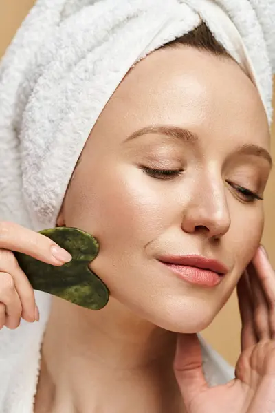 Une femme avec une serviette enroulée autour de sa tête pose avec une gua sha verte sur son visage, mettant en valeur sa beauté naturelle. — Photo de stock