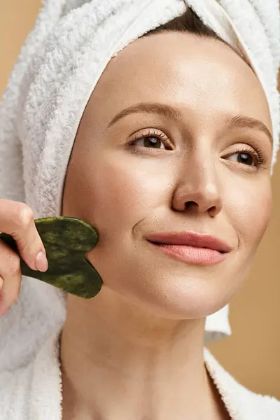 A woman with a towel on her head delicately holds a green face roller, exuding a sense of tranquility and connection with nature. — Stock Photo