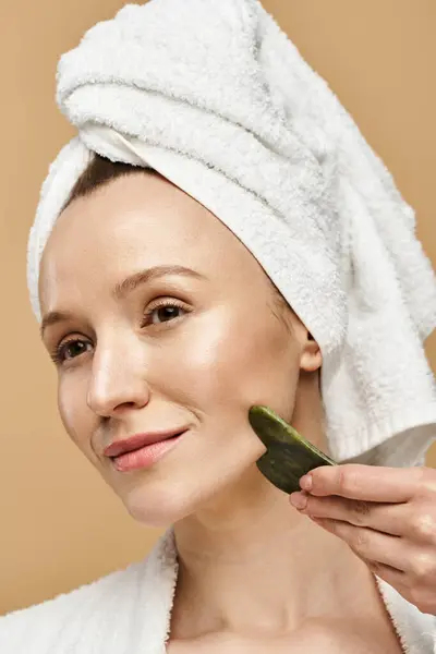 A stunning woman with a towel wrapped around her head holds face roller in her hand, exuding relaxation and refreshment. — Stock Photo