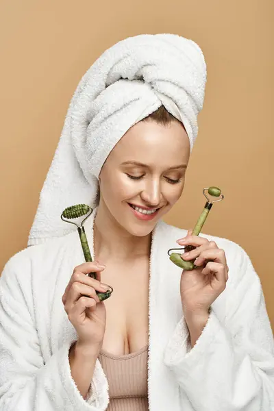 An attractive woman with a towel wrapped around her head holding two face rollers, showcasing her natural beauty. — Stock Photo