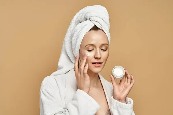 A graceful woman with a towel wrapped around her head delicately holds a jar of cream, showcasing her natural beauty routine. — Stock Photo