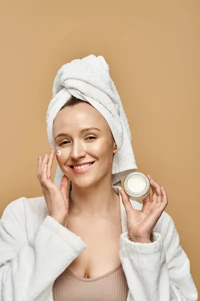 Una mujer serena con una toalla en la cabeza sostiene con gracia un frasco de crema, enfatizando su belleza natural y su rutina de cuidado personal. - foto de stock