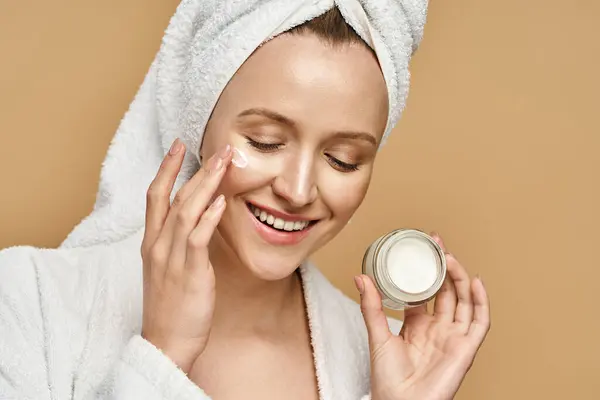 Una donna attraente con un asciugamano sulla testa che si applica crema sul viso. — Foto stock