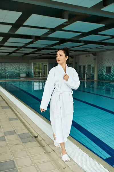 Une femme brune en peignoir se tient élégamment à côté d'une piscine intérieure dans un spa luxueux. — Photo de stock