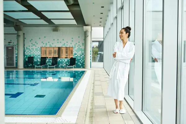 Молода брюнетка в халаті стоїть витончено поруч з критим басейном у спа-центрі. — стокове фото