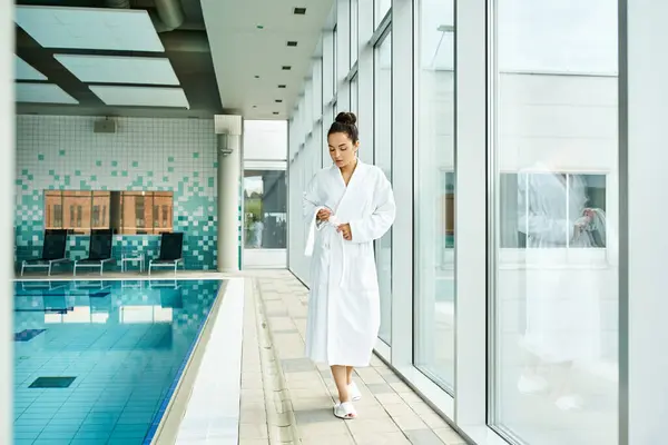 A young, beautiful brunette woman in a bathrobe standing beside an indoor swimming pool. — Stock Photo