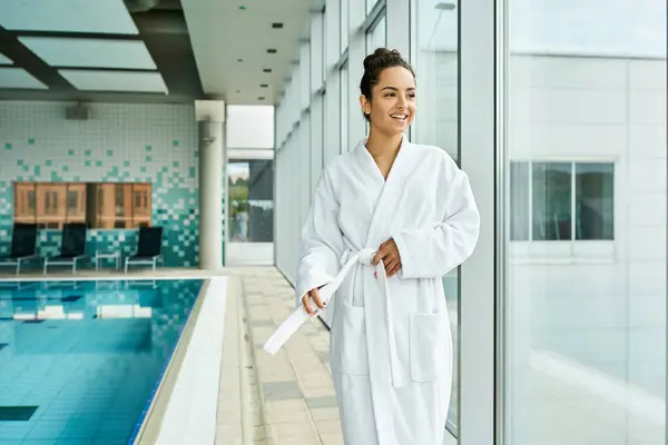 A young, beautiful brunette woman in a bathrobe standing next to an indoor swimming pool. — Stock Photo