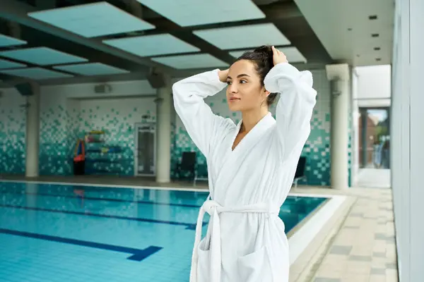 A young, beautiful woman in a bathrobe stands gracefully beside an inviting swimming pool in an indoor spa setting. — Stock Photo