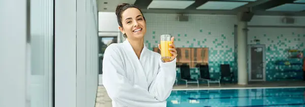 A brunette woman in a luxurious bathrobe leisurely sips orange juice by an indoor spa with a swimming pool. — Stock Photo