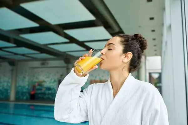 Eine junge, schöne brünette Frau genießt luxuriös ein erfrischendes Glas Orangensaft in einem Indoor-Spa am Swimmingpool. — Stockfoto