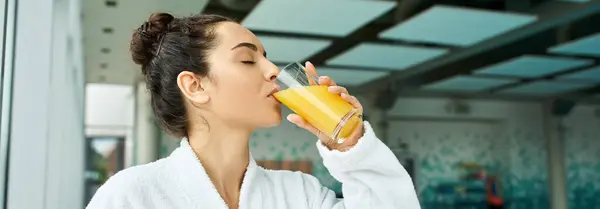 Una joven y hermosa morena saboreando un vaso de zumo de naranja refrescante en un spa interior con piscina. - foto de stock