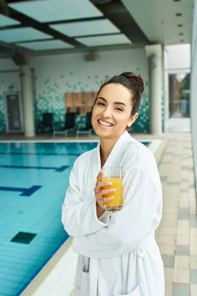 A young brunette woman relaxes in an indoor spa, savoring a glass of orange juice in her bathrobe by the swimming pool. — Stock Photo