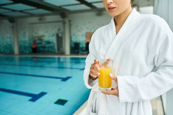 Una joven y hermosa morena en un albornoz se relaja junto a una piscina, sosteniendo un vaso de jugo de naranja. - foto de stock