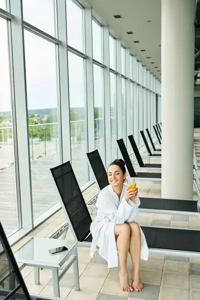 A young, beautiful brunette woman sits serenely on a bench in an indoor spa with a swimming pool, clad in a luxurious bathrobe. — Stock Photo