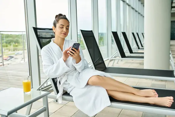 Молода брюнетка в халаті відпочиває в критому спа-центрі, використовуючи мобільний телефон. — стокове фото