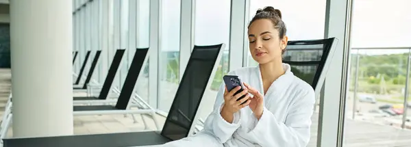 Una giovane donna bruna seduta su una sedia, affascinata dallo schermo del suo cellulare, in una spa coperta con piscina. — Foto stock