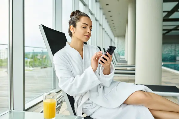 A young, brunette woman is seated in a luxurious bathrobe, engrossed in her cell phone in an indoor spa with a swimming pool. — Stock Photo