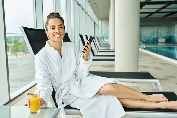 Jeune femme brune profitant d'un moment paisible dans une chaise longue au bord d'une piscine, absorbée dans son téléphone portable. — Photo de stock