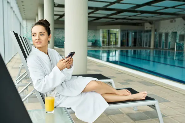 A young, beautiful brunette woman in a bathrobe is sitting on a lounge chair by an indoor swimming pool. — Stock Photo