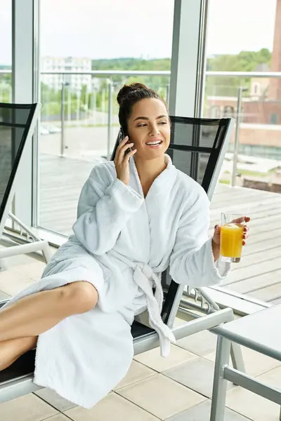 A young brunette woman is sitting in a lounge chair by an indoor spa pool, engaged in a phone conversation. — Stock Photo