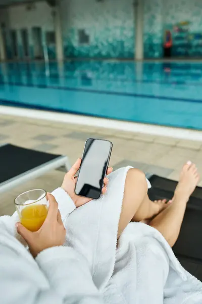 Una joven morena tendida junto a una piscina cubierta, en paz usando su teléfono celular. - foto de stock