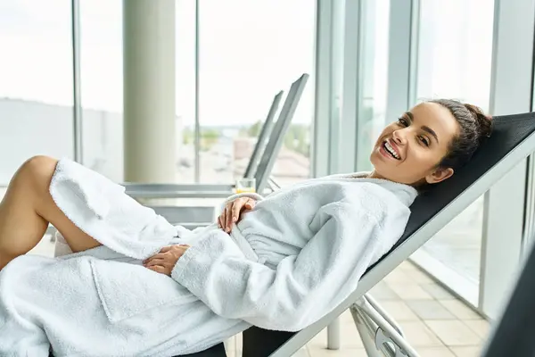 A young, beautiful brunette woman in a bathrobe sits peacefully in a lounge chair indoors at a spa with a swimming pool. — Stock Photo